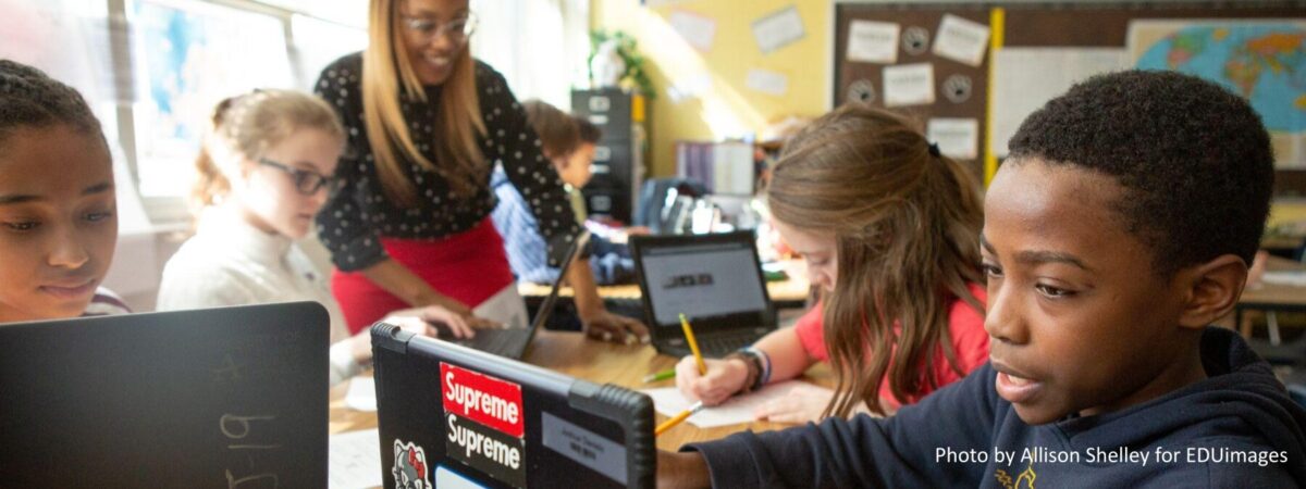 A photograph showing a teacher standing over a table, assisting a diverse group of elementary school students working on individual Chromebooks.