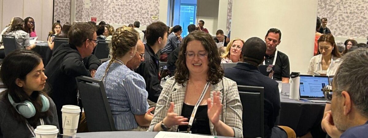 A conference room filled with roundtables. The primary focus is on one table, with a woman speaking to the group.