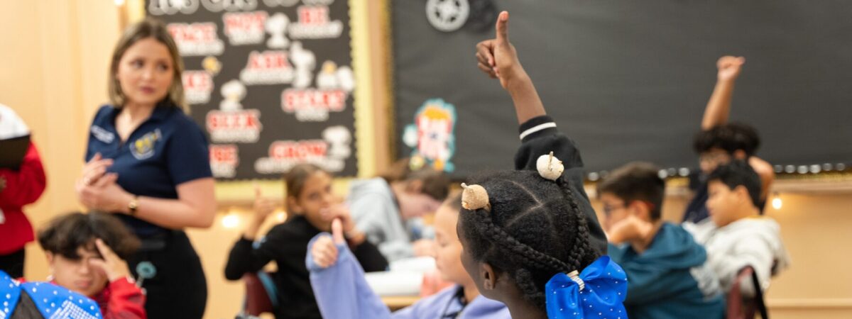 Students raise their hands with hands up or thumbs up during a classroom lesson, while a teacher stands near a bulletin board at the front of the room.