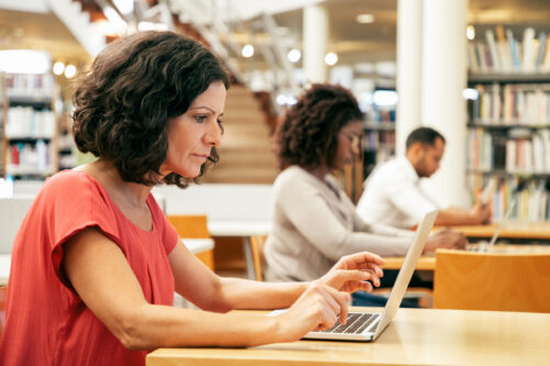 Serious focused middle aged woman working on research in public library. Adult students sitting at desks and using laptops. Academic research concept