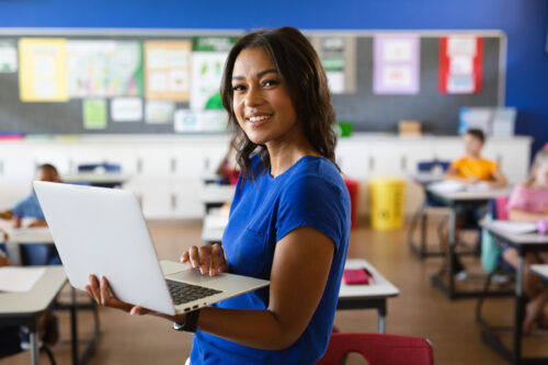 A smiling woman in a blue T-shirt holds an open laptop while standing in a classroom. Students sit at desks behind her, and a bulletin board with colorful posters hangs on the wall.