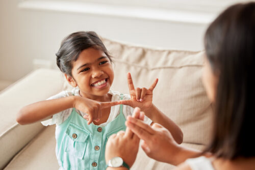 A smiling girl in a mint-green jumper sits on a beige couch and uses her hands to sign to an adult, who is shown from behind and is also signing.