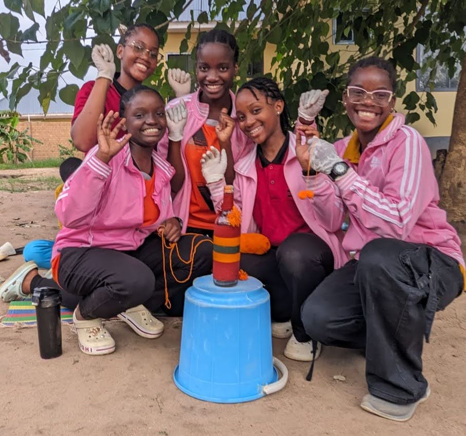 Five students in pink jackets pose crouched around their design