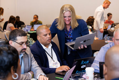 A participant holding a laptop leans in to help attendees working on laptops at a Leader Summit table session.