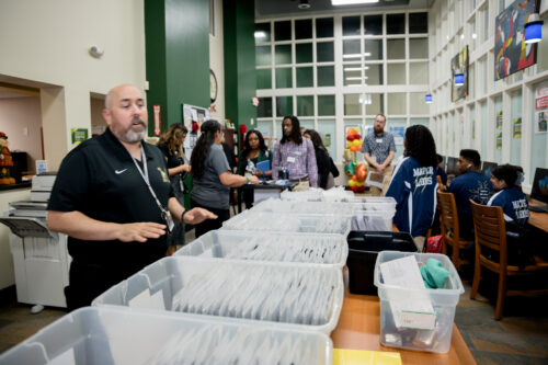 A staff member explains bins of bagged tablets to visitors in a school library as students work at nearby computers.