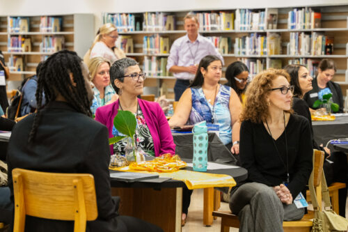 A group of teachers sit at tables and stand in a library while watching a speaker.