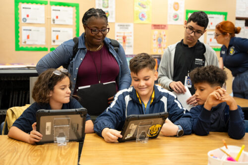 Middle school students use tablets at a classroom table as a smiling teacher and a Verizon Innovative Learning coach look on.