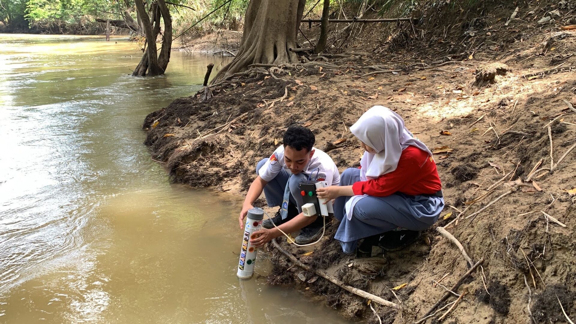 Two students crouch over a river bank with the FlowSafe device.