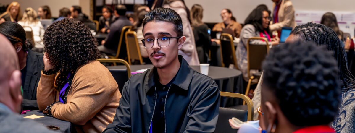 An image of a student in a gray suit wearing glasses who is talking to adults while at a conference.