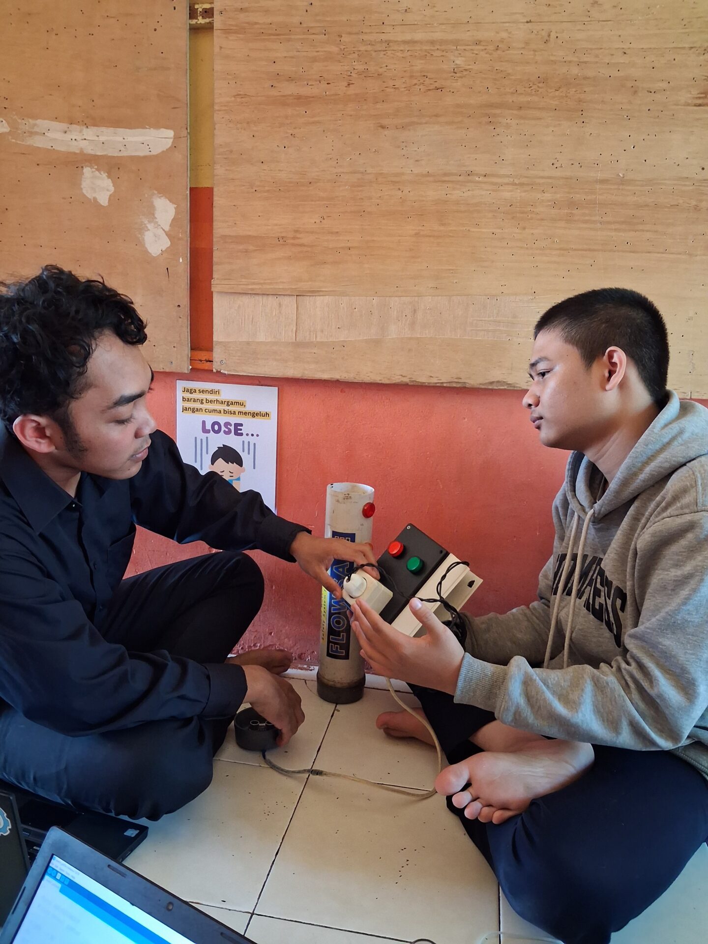 Two students sit on a tiled floor while holding their devices.