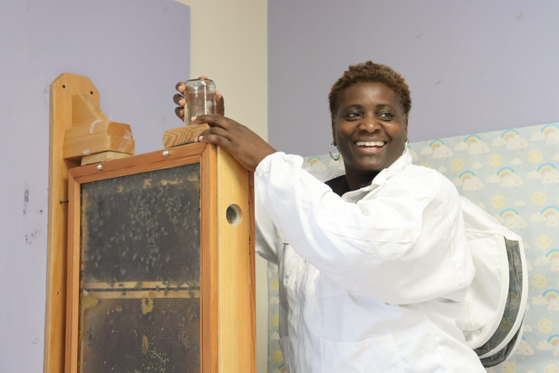A woman is smiling in a white beekeeping suit with the hood down while she rests her hand on top of an observation beehive device with bees visible on the honeycomb inside. 