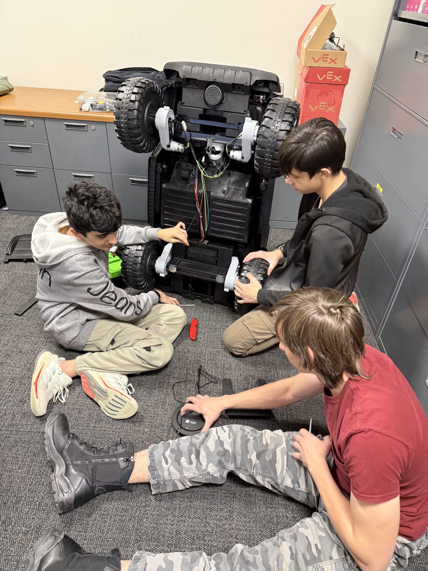 Three students sit on the floor while working on a toy car.