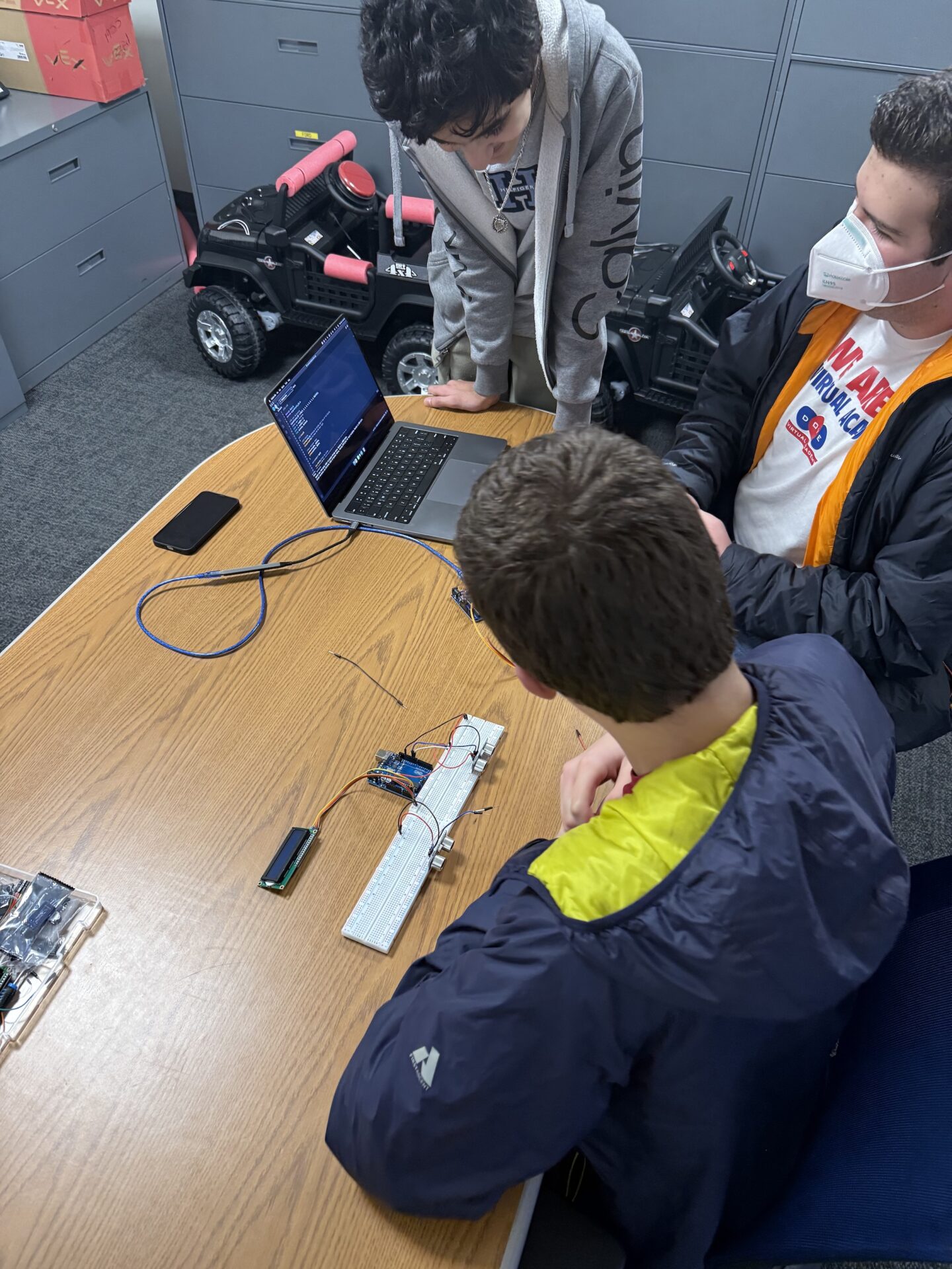 Three students gather at a table with a laptop and programming devices.