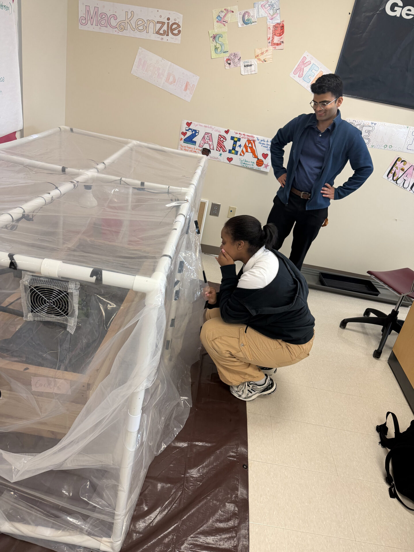 A teen crouches to label the plastic covering of a homemade PVC-frame greenhouse with a marker while a teacher stands nearby watching a smiling, in a classroom decorated with student name art.