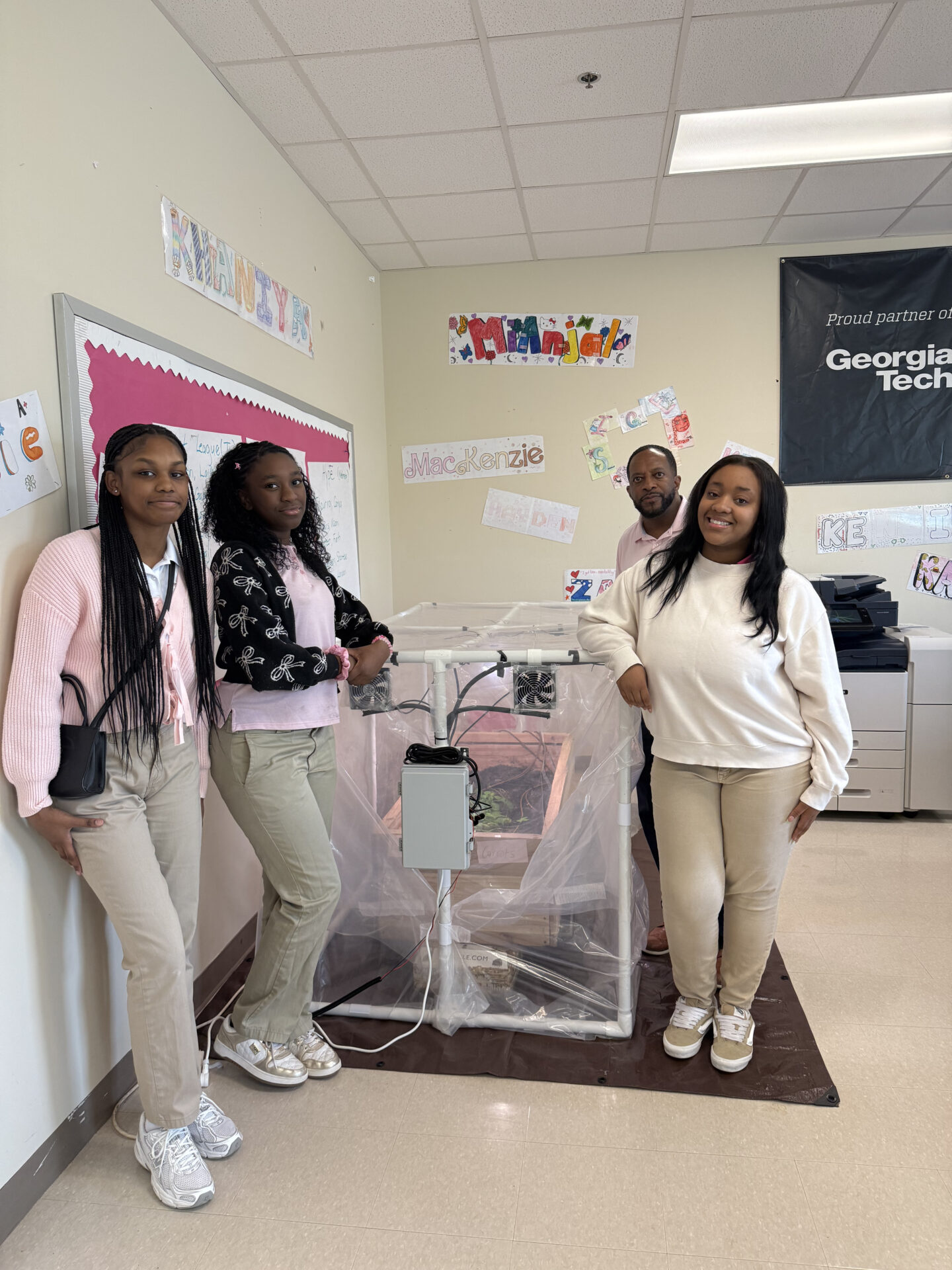 Three teens and a man stand in a classroom around a homemade PVC-frame greenhouse enclosed in clear plastic, fitted with fans and wiring, with a "Georgia Tech" banner and student name decorations on the walls behind them. 