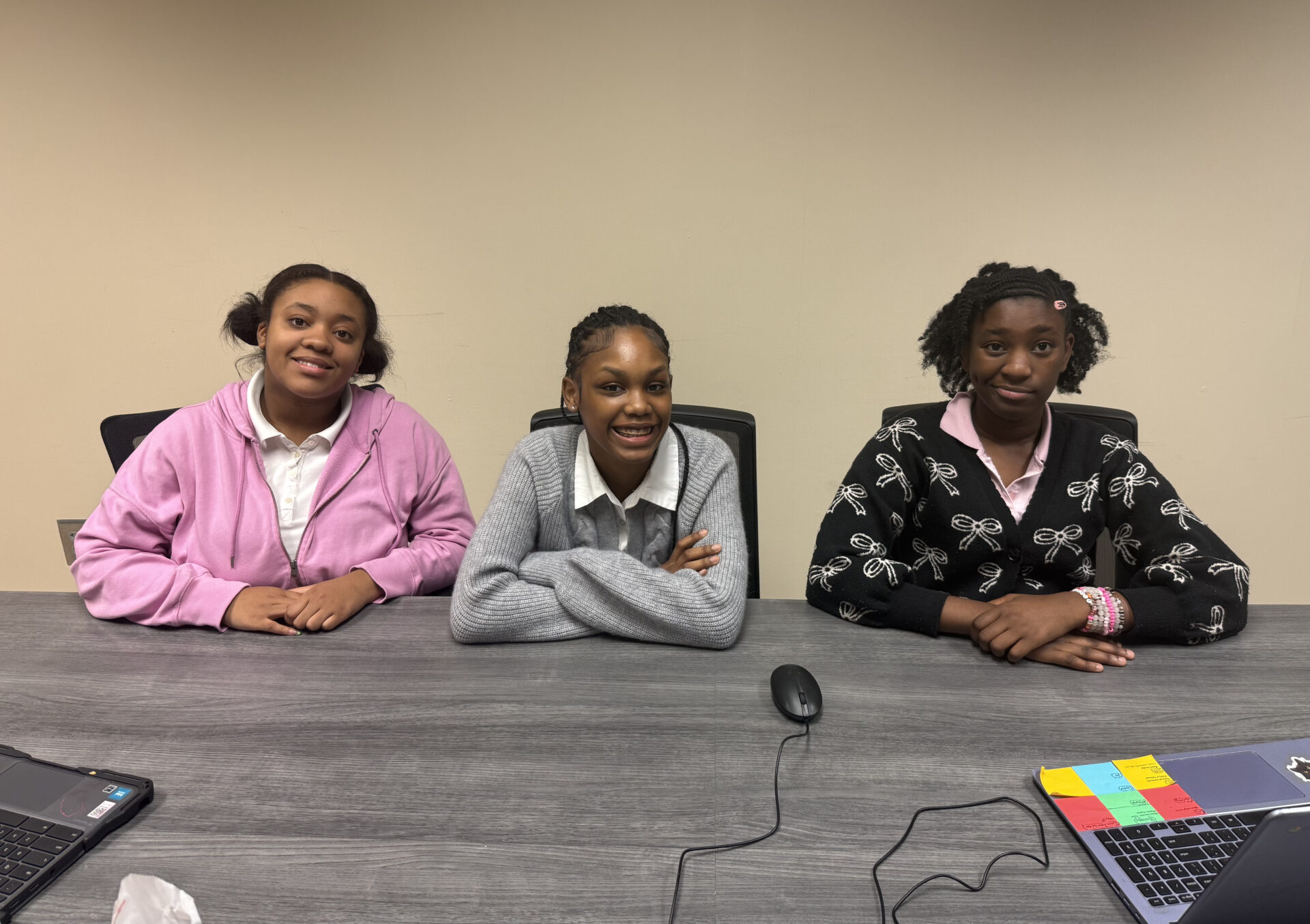 Three teens sit smiling at a conference table with laptops, mouse, and colorful sticky notes in front of them. 