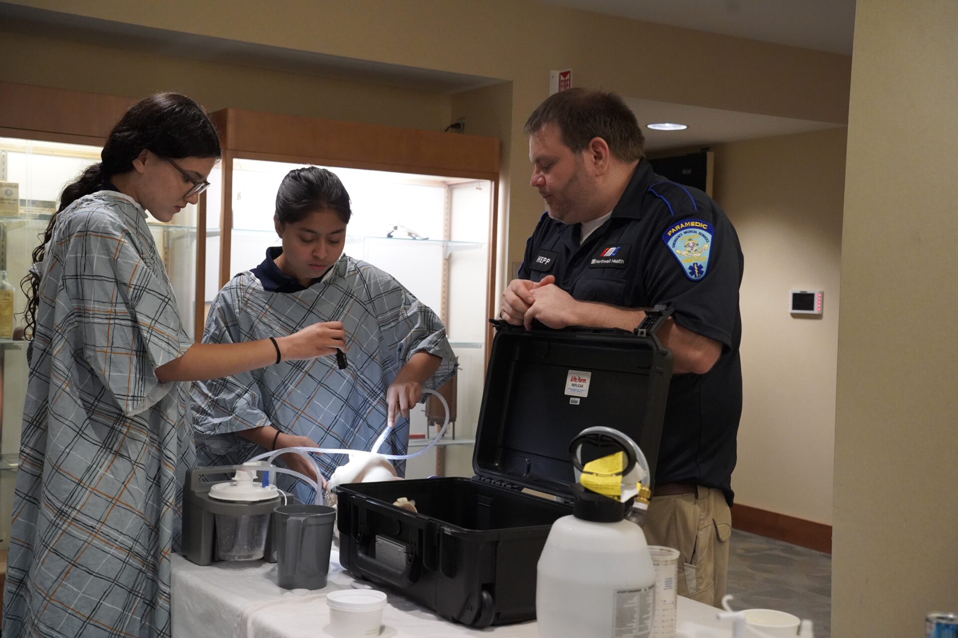Two high school students wearing medical gowns work on equipment with an EMT officer. 
