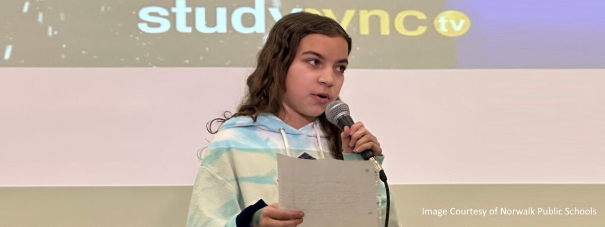 A middle school student stands on a stage holding a microphone and reading from a paper.