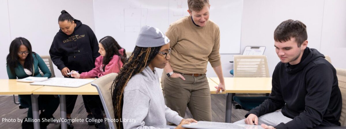 Instructor discussing a math problem with two students over a whiteboard as another small group works together in the background, in a classroom on a college campus.