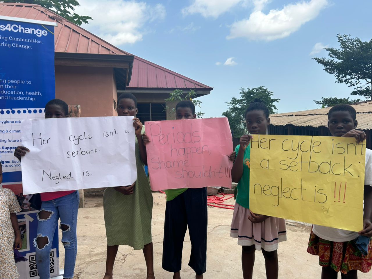 Students hold up signs that say "her cycle isn't a setback. Neglect is," and "periods happen, shame shouldn't!!!"