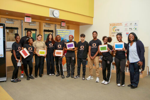 A student tech team of student leaders wearing matching t-shirts smile while holding posters.