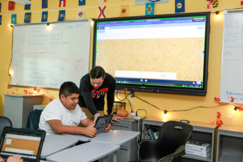 A teacher helps a student on a tablet while there is a lesson on a large screen.