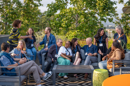 Smiling conference attendees with name badges chat and laugh together on sofas on a leafy, sunlit rooftop terrace.
