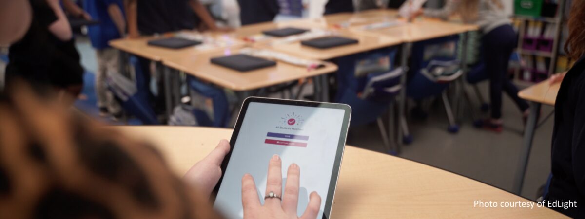 A teacher holds a tablet with an app that says All Students Matched on screen in a classroom with students
