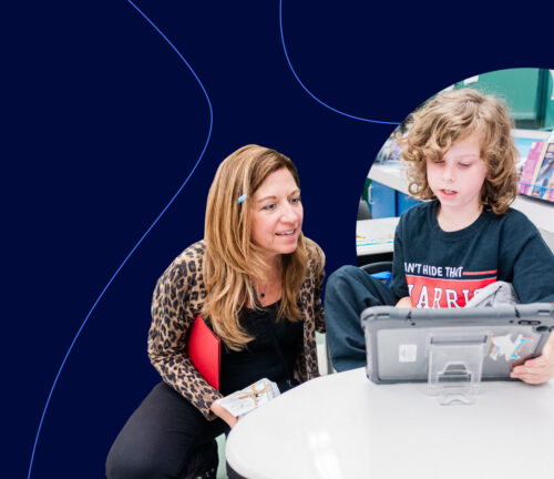 A teacher crouches next to a young student as they work on a tablet