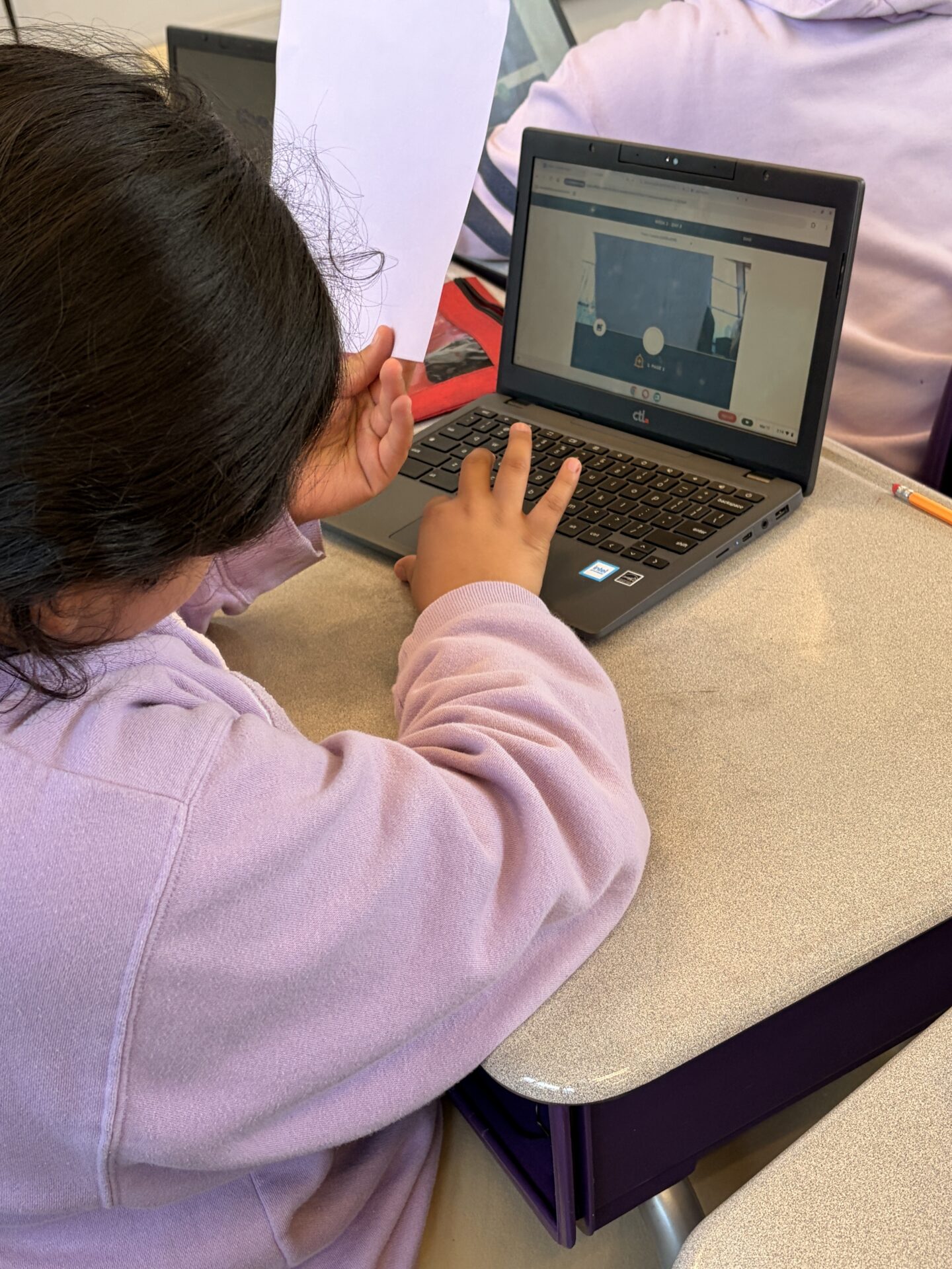 An elementary school students types on a laptop during an exercise and holds up a piece of paper. 
