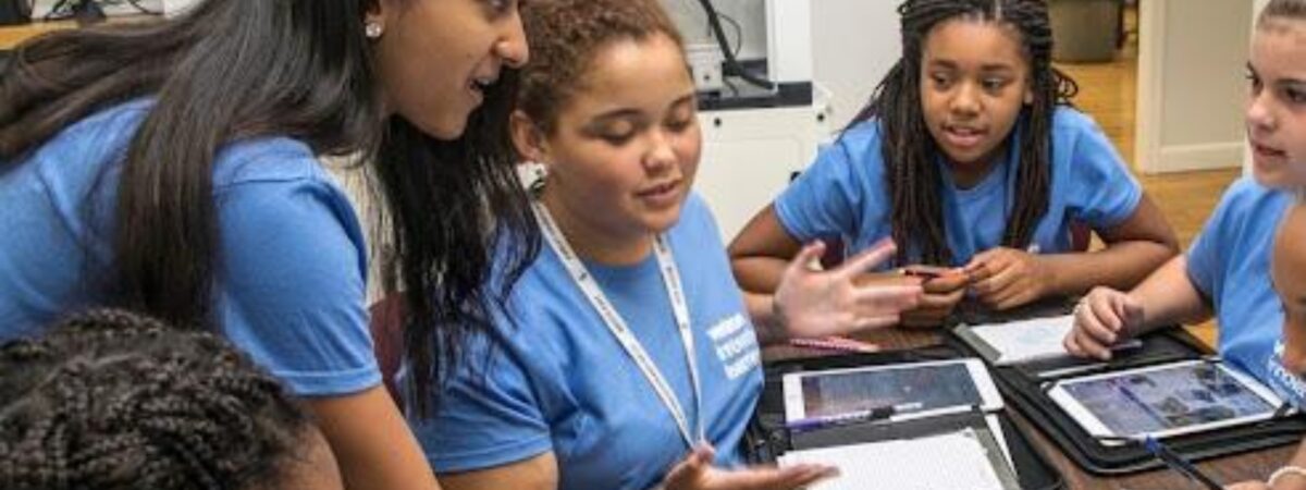 A young woman in a blue Verizon Innovative Learning T-shirt leans in to talk with a group of girls in matching blue T-shirts gathered around a table with tablets and worksheets. A science lab with a fume hood is in the background.