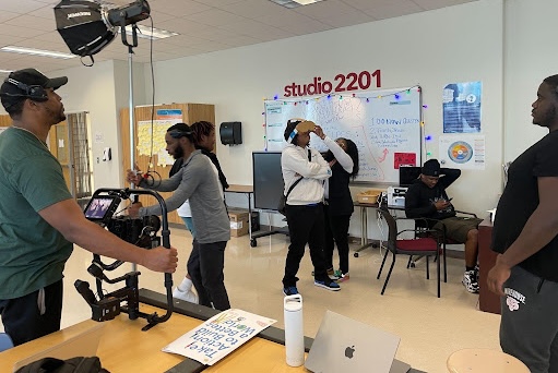 Students film a video in a room with a "studio 2201" sign on the wall. One student in headphones operates a camera on a stabilizer rig while others prepare a scene near a whiteboard, with a studio light overhead and a laptop on a table in the foreground.