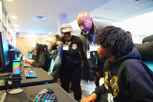 A student in a hoodie sits at a computer with a backlit keyboard while two smiling adults wearing name badges lean in to watch. Other students sit at a row of computer stations in the background.