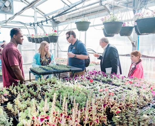 Five people gather among tables of seedlings and blooming flowers in a greenhouse with hanging flower baskets overhead. A student holds a tray of seedlings while two adults, one in a suit, look on and talk, joined by a young man and a girl in a pink jacket.