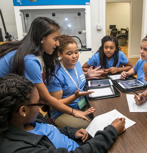 A young woman in a blue T-shirt that says Verizon Innovative Learning leans in to talk with a group of girls in matching blue T-shirts gathered around a table with tablets and worksheets. A science lab with a fume hood is in the background.