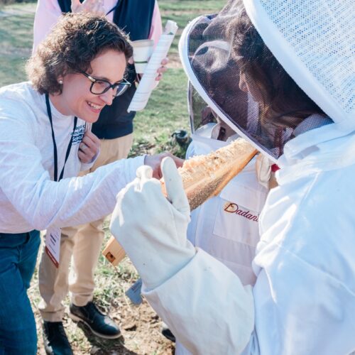 A young student in a bee suit holds a honeycomb in front of an adult