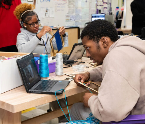 Two students work with yarn and follow instructions on their laptops.