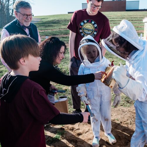 A young student in a bee suit holds a honeycomb in front of an adult